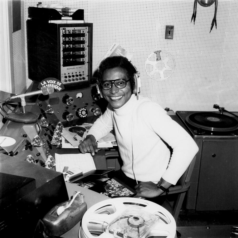 Collinet in a white turtleneck, sitting at a radio broadcasting panel with headphones on, smiling at the camera