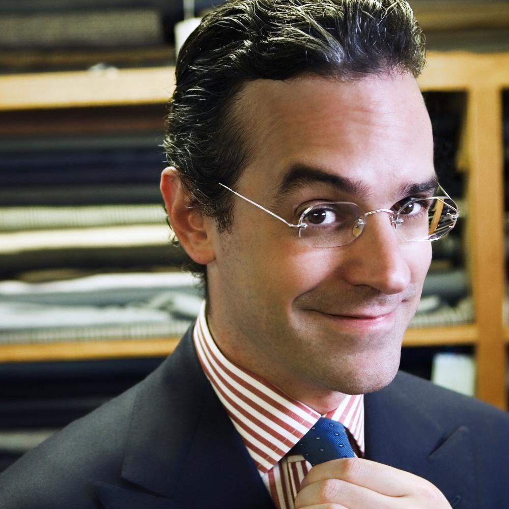 Anton, wearing rimless glasses and hair slicked back, adjusts his blue tie, worn with a red and white striped shirt and black blazer