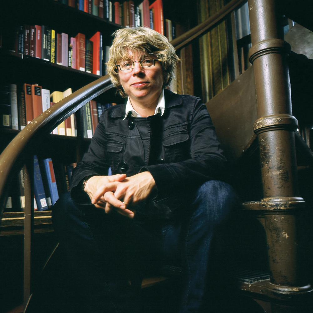 Lepore sits on a step of a winding brown wood staircase, surrounded by shelves of books