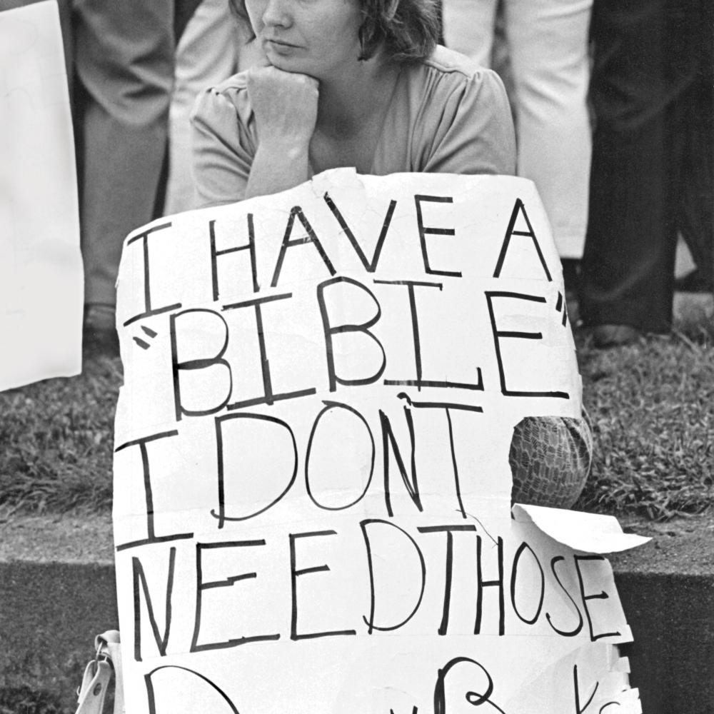 Black and white photo of a woman sitting on a curb holding a sign of protest with her chin on her fist.