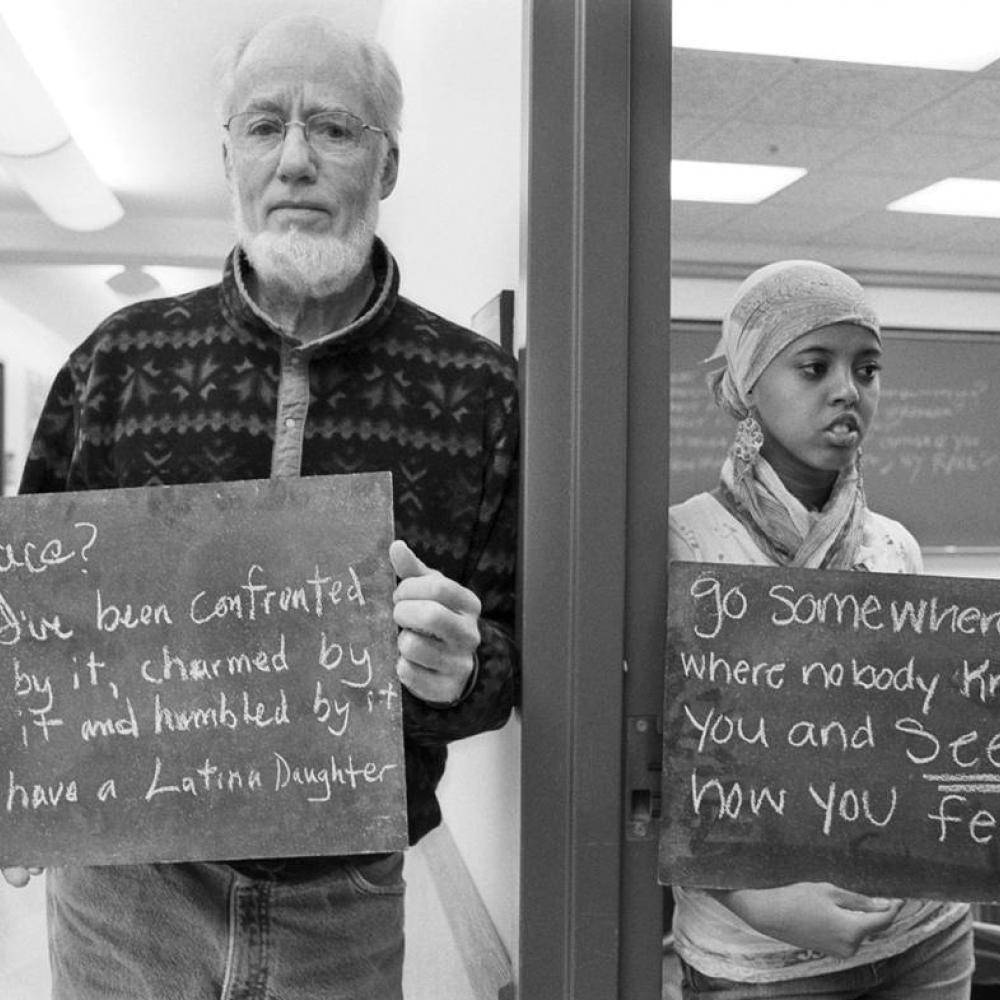 Black and white photo of two individuals holding signs that have provocative statements about race on them.