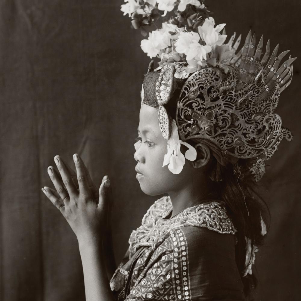 Side view of a young balinese dancer, palms clasped together, wearing a tall floral headdress and embroidered dress