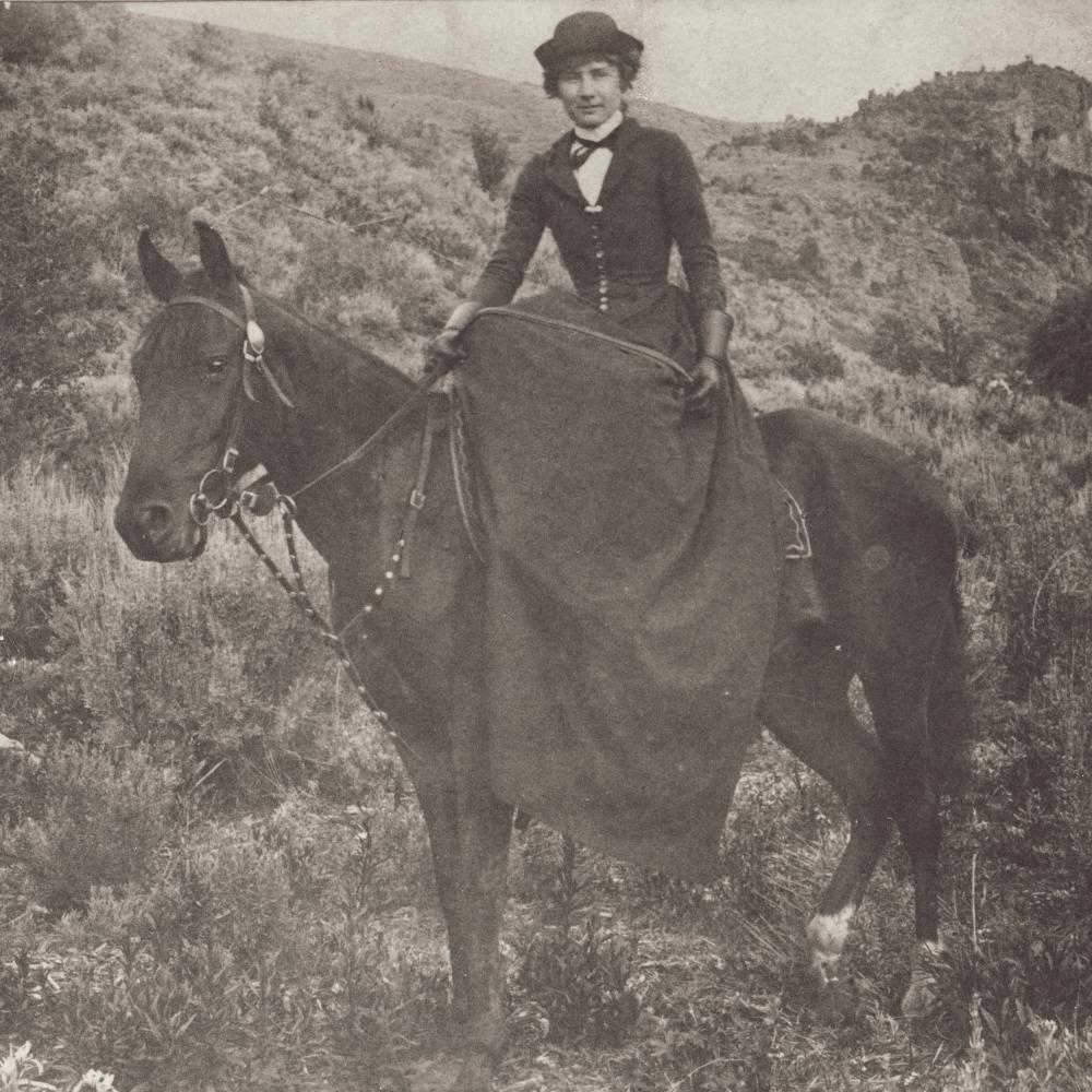 Black and white photo of a woman riding a black horse, wearing a full dress and hat.