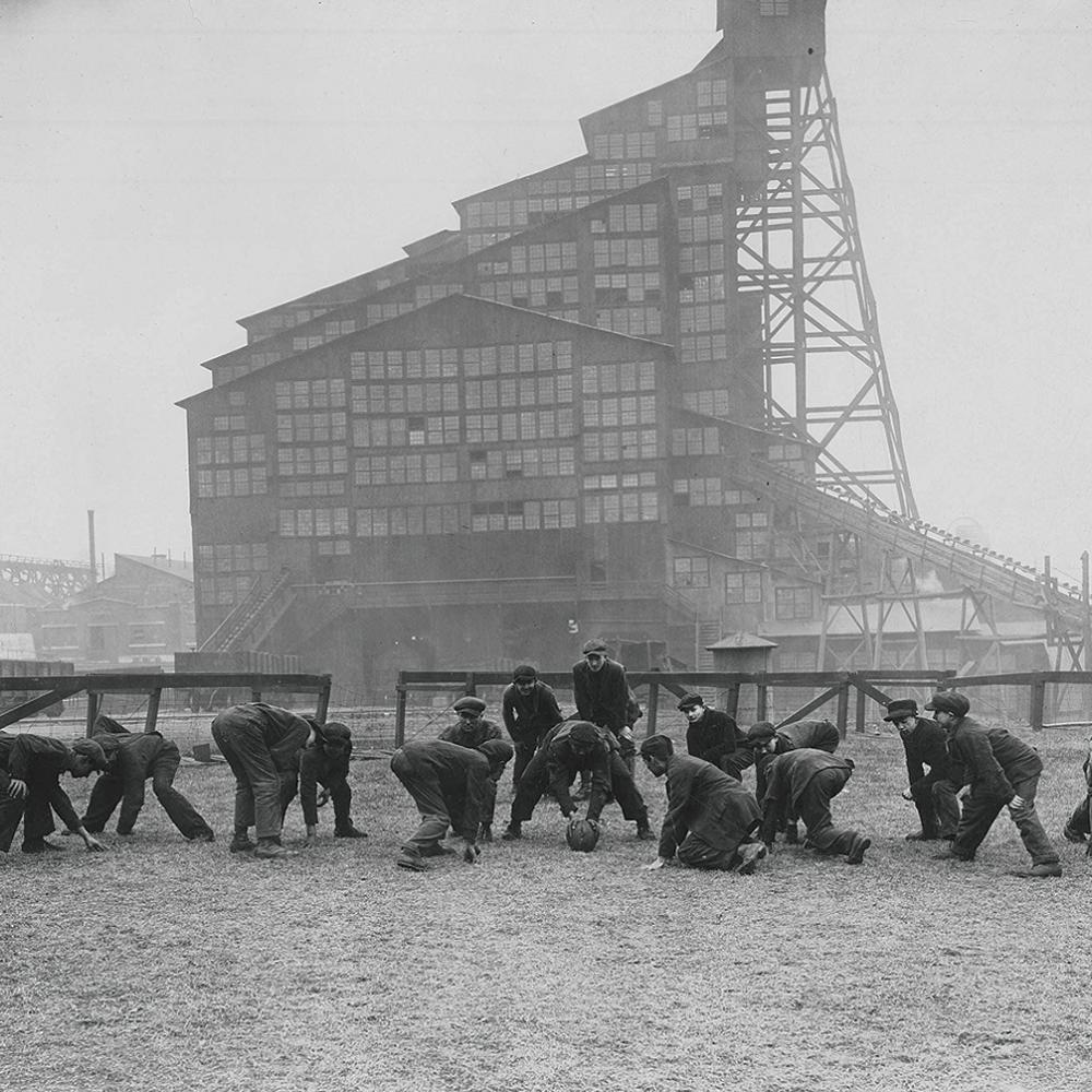 Black and white photo of boys playing football with a mining facility in the background.