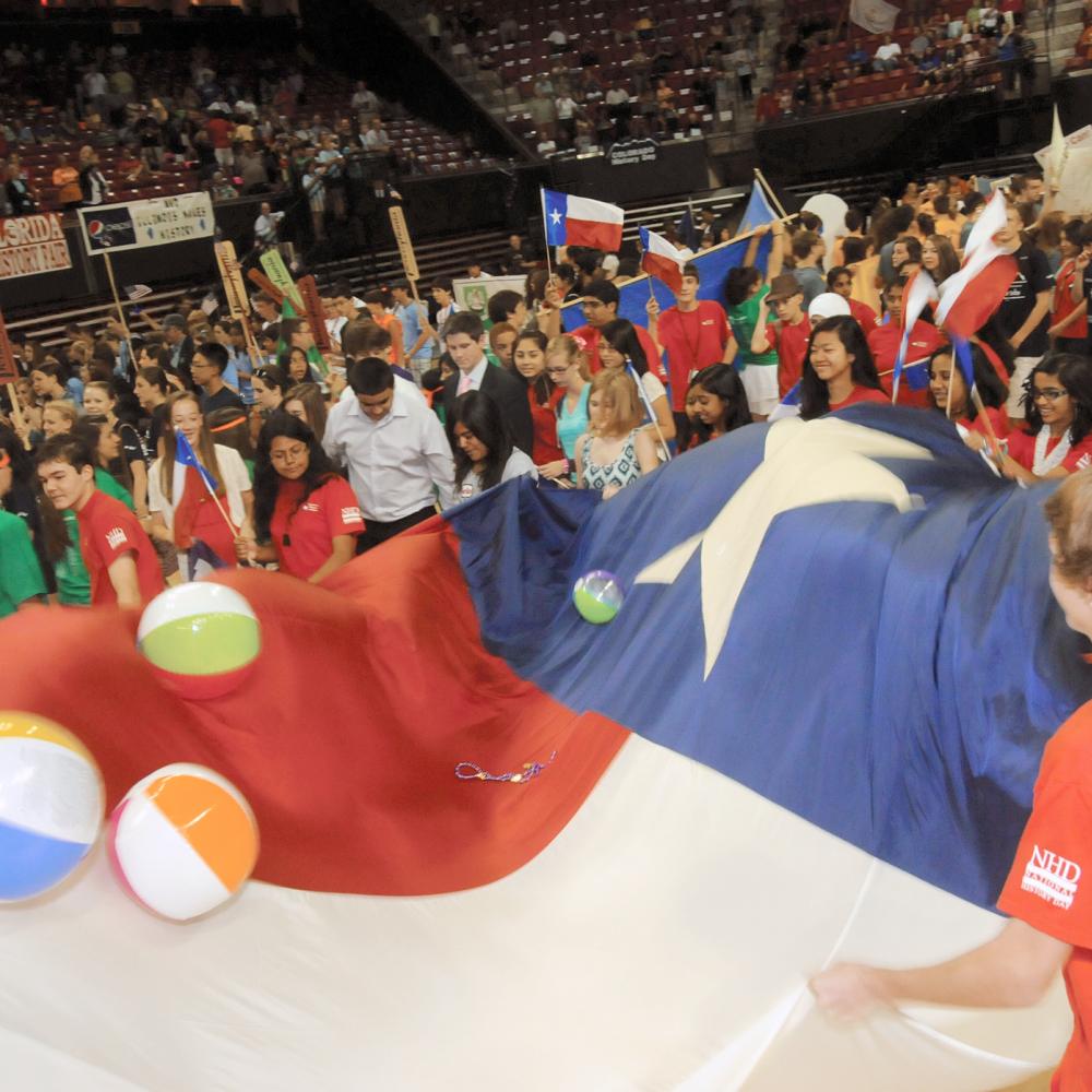 Color photo of a crowd of young students flopping inflatable balls around on top of a large Texas flag.