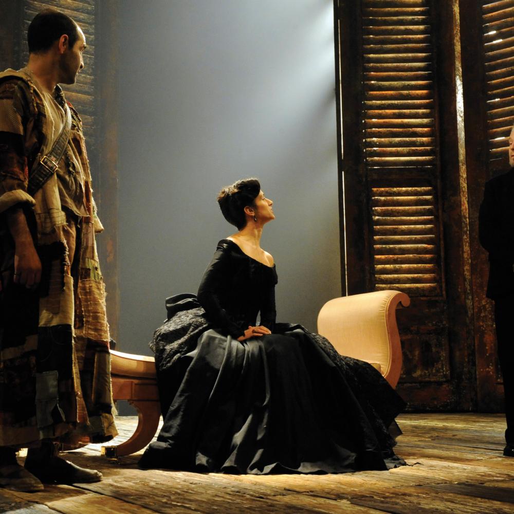 Female actor in a black velvet dress, seated, looks defiantly at the male actor in a dark suit, during a scene of Twelfth Night