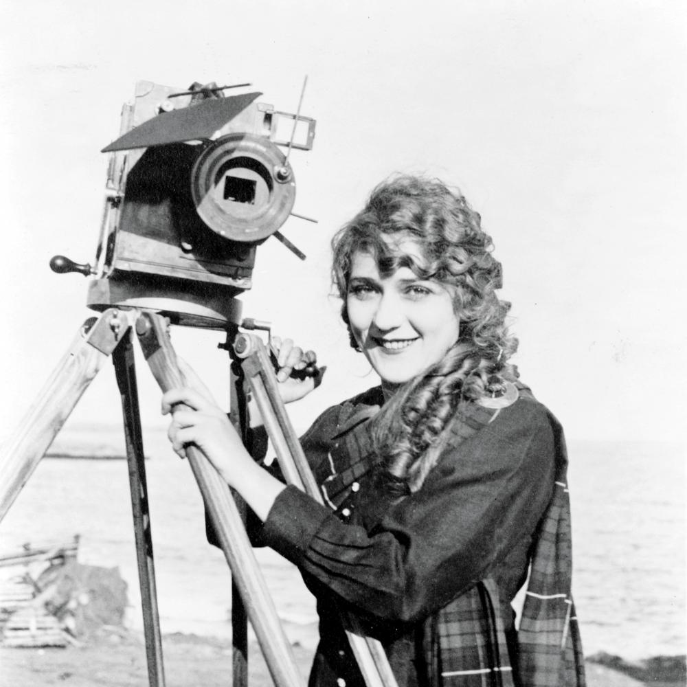 Pickford in a plaid coat, standing with one of her cameras and smiling at the camera