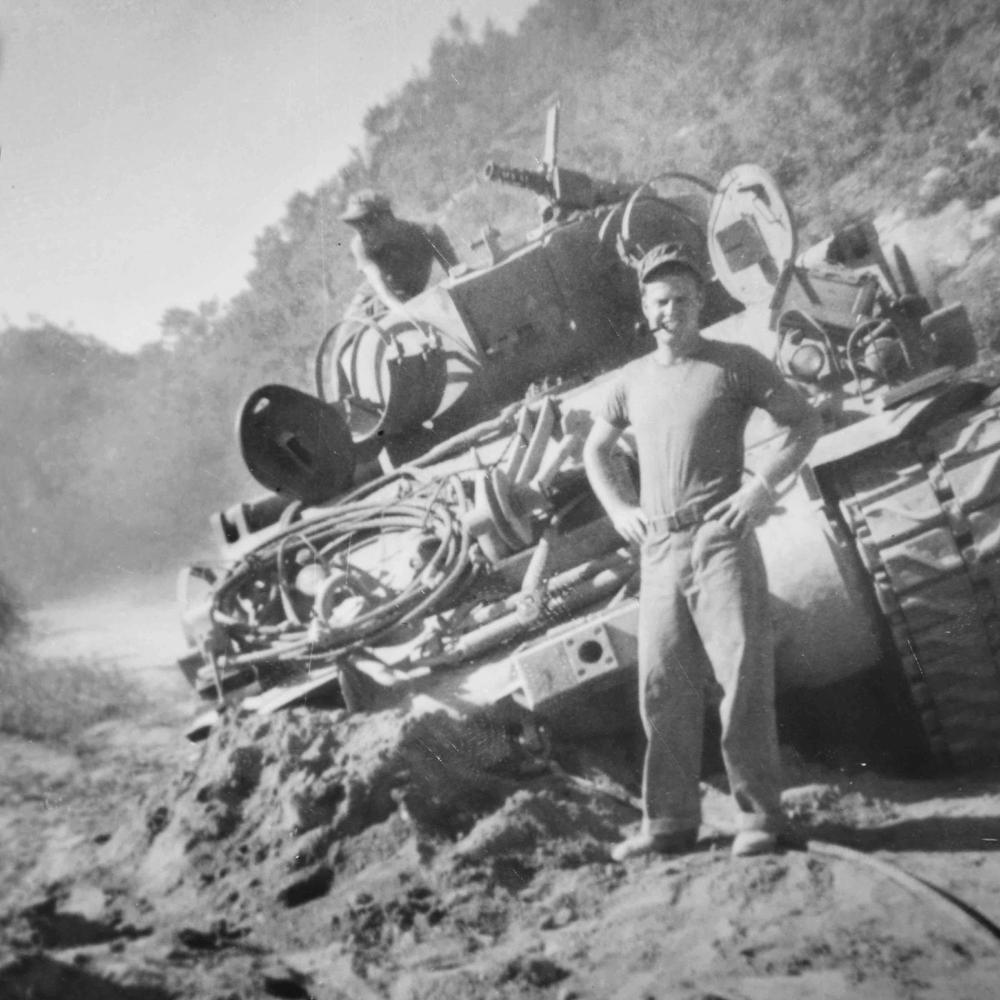 McInroe stands with his hands on his hips, pipe in his mouth, in front of his tank