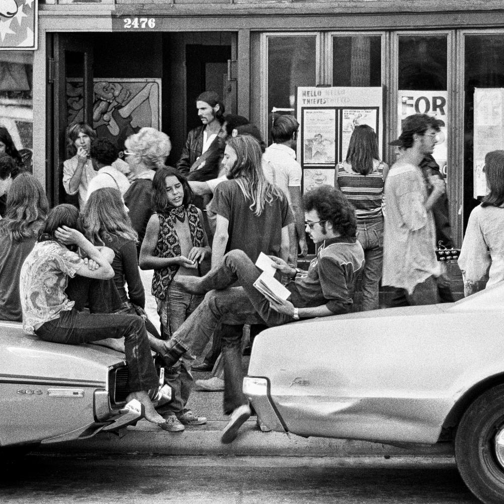Black and white photograph of people standing around and lounging on cars