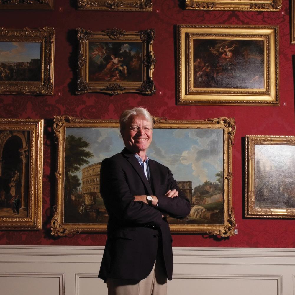 Color photo of Gary Vikan standing before a wall of paintings. He wears a black blazer and khaki dress pants.