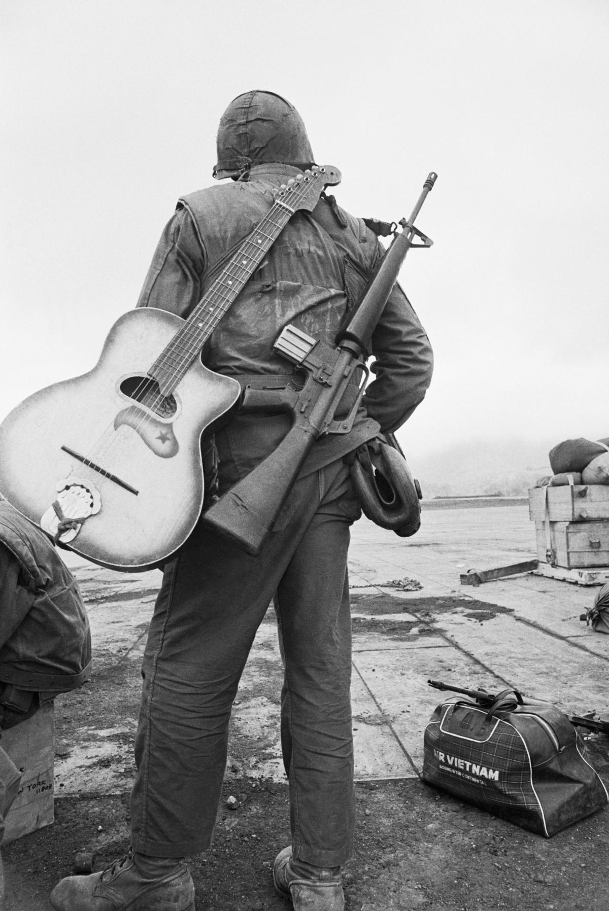 Black and white photo of a soldier with an M16 and a guitar slung over his back.