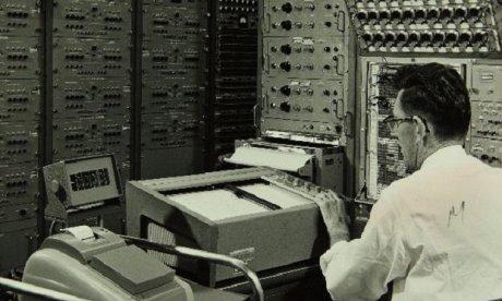 Black and white photo of a man sitting before a display and surrounded by large early computers.