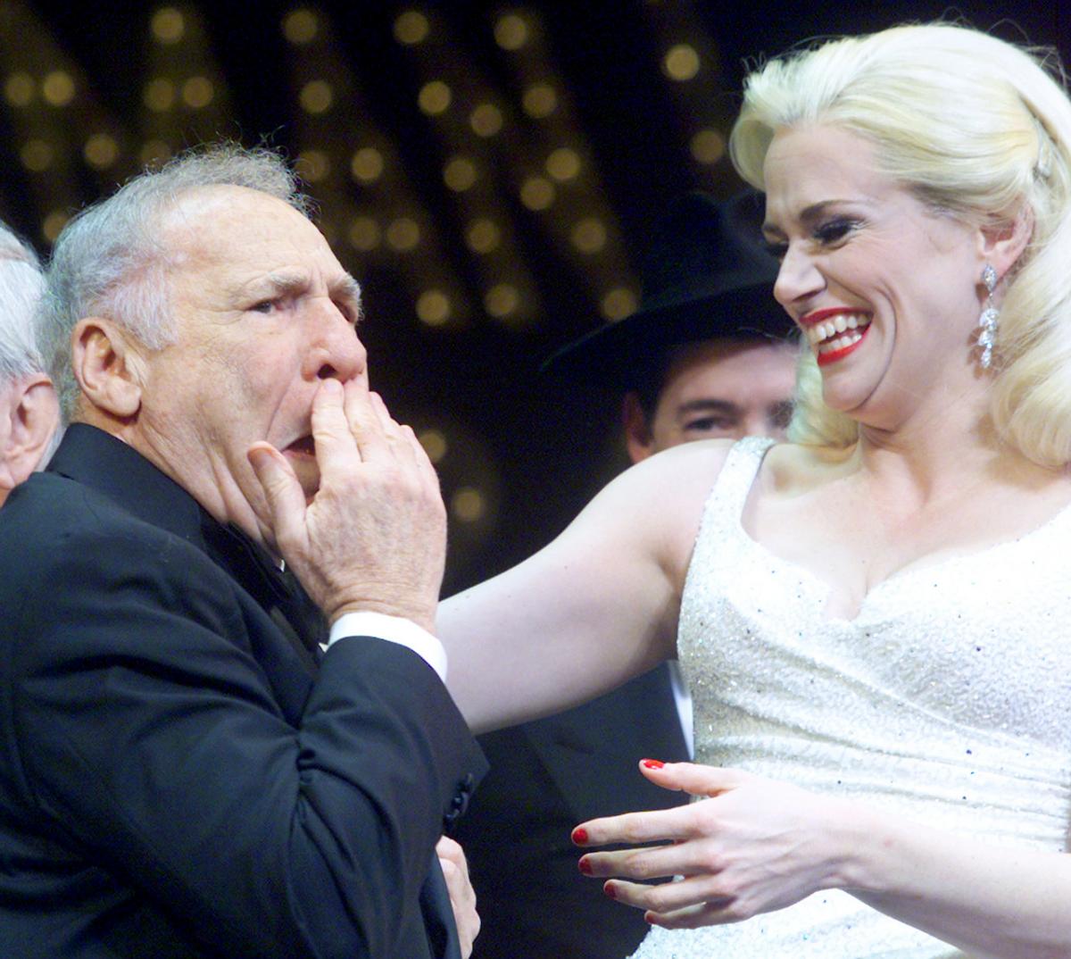 Brooks holds his hand to his face in surprise as a blonde actress in a white dress reaches out to embrace him