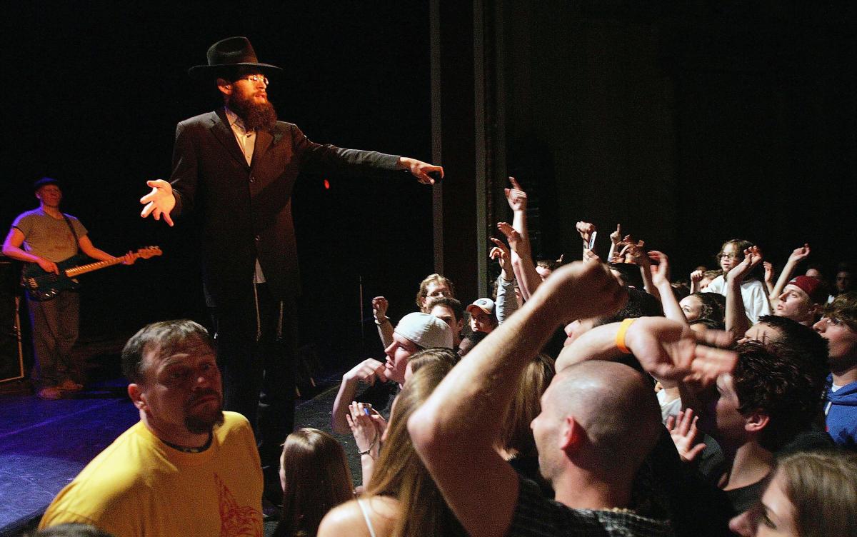 Matisyahu on stage, in a black suit and black hat, points to a crowd of concert goers while performing