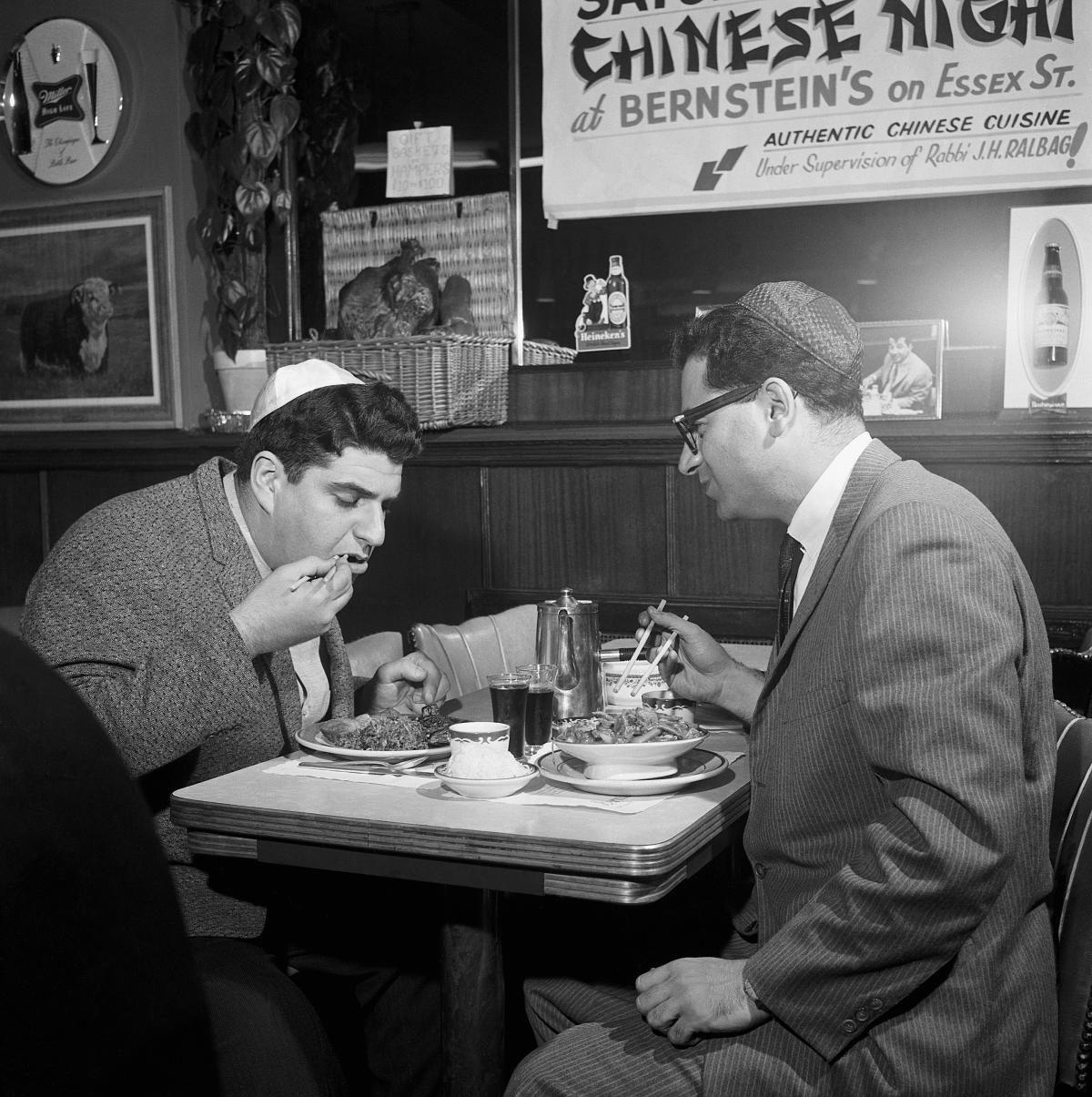 Two men wearing yarmulkes sit at a diner table, eating chinese food with chopsticks