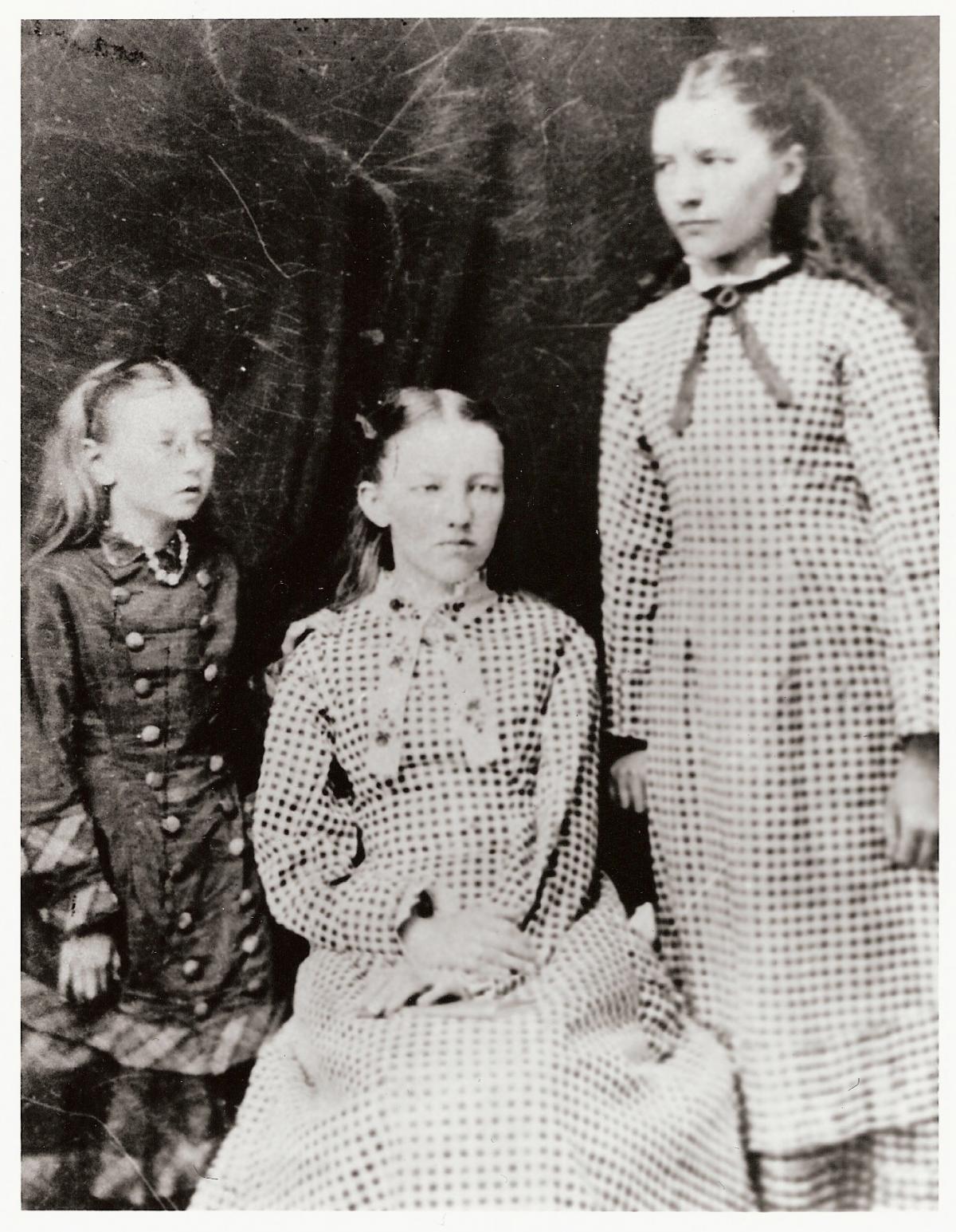 Black and white photograph of three young girls, two seated and one standing, in checkered and buttoned dresses