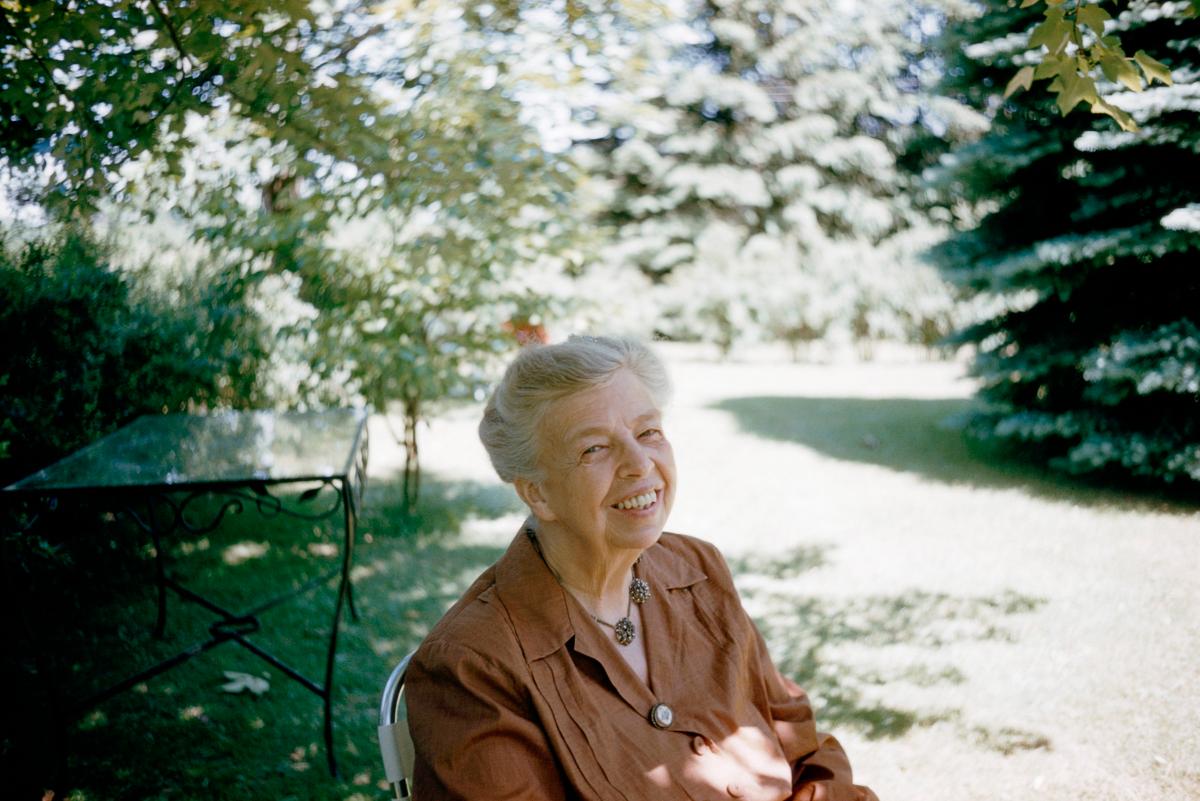 Eleanor Roosevelt, in a brown blouse, sitting in a green backyard under dappled sunlight