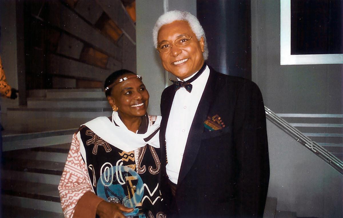 Collinet in a black tuxedo smiles at the camera, next to Makeba, wearing a thing beaded band around her forehead, and smiles at him