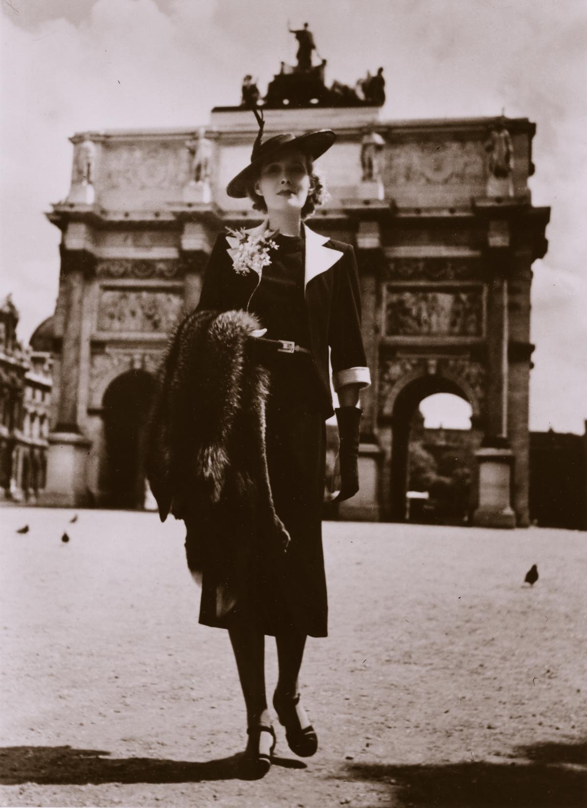 Bedwell, wearing black dress, pumps, black jacket with white collar, and a flat black hat with a feather, carrying a fur stole, in front of the stone Arc