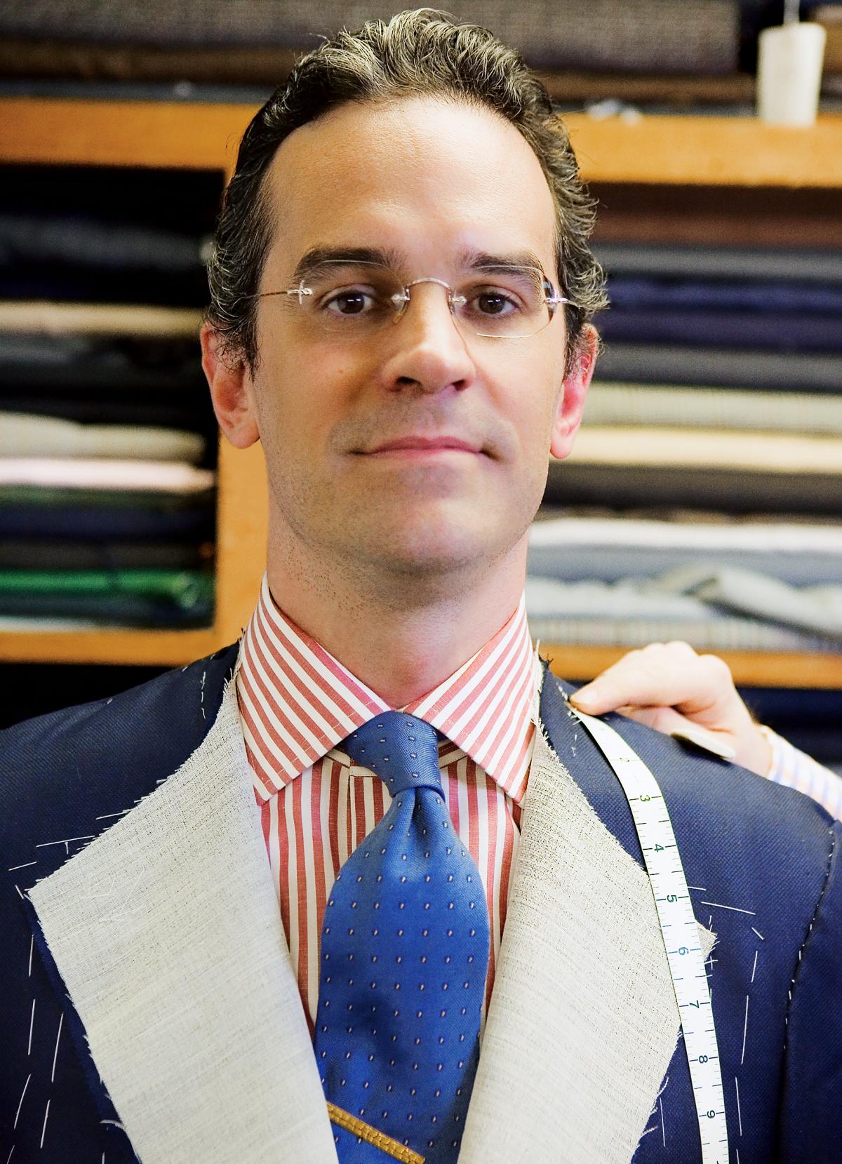 Anton looks directly at the camera while getting measured for a new, navy blue suit, which he wears over a red and white striped shirt and cobalt blue tie