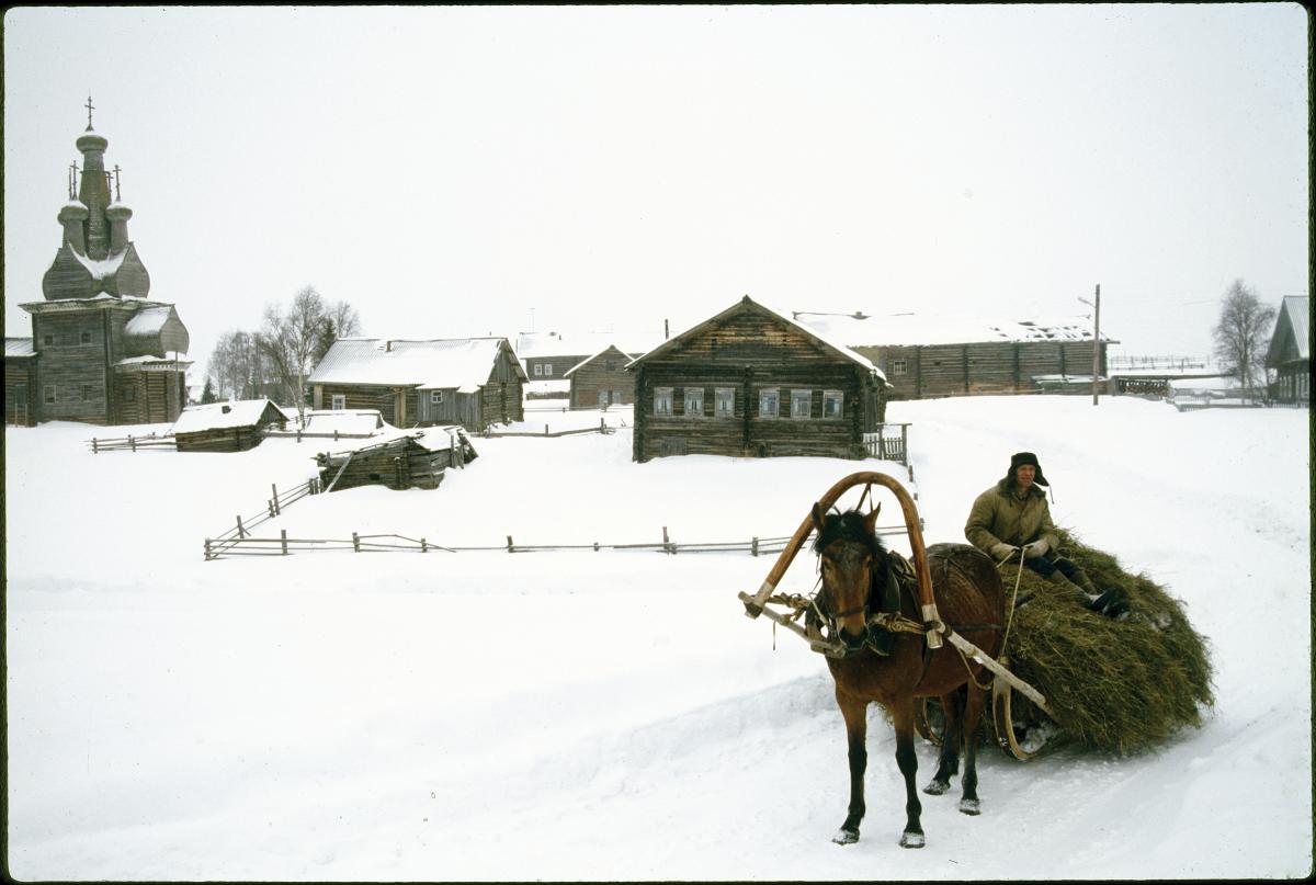 Photograph of a snowy landscape, man in cart being pulled by horse, log house in background