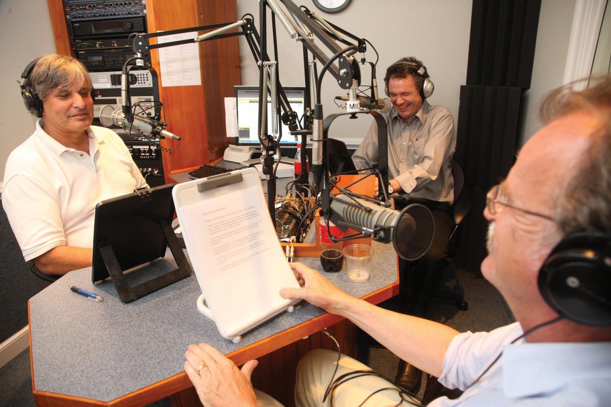 The three hosts look at clipboards of notes while recording a session, wearing headsets and sitting in front of their mics