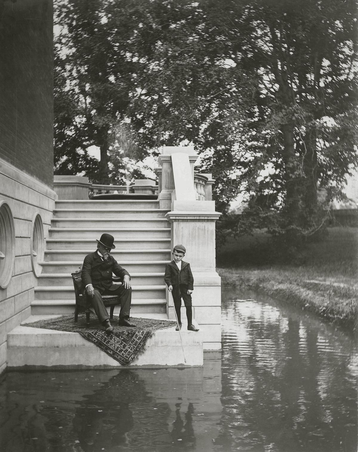 Wallace, in a bowler hat and dark suit, sits on a chair and looks down into the moat, while his grandson dips a short pole into the water