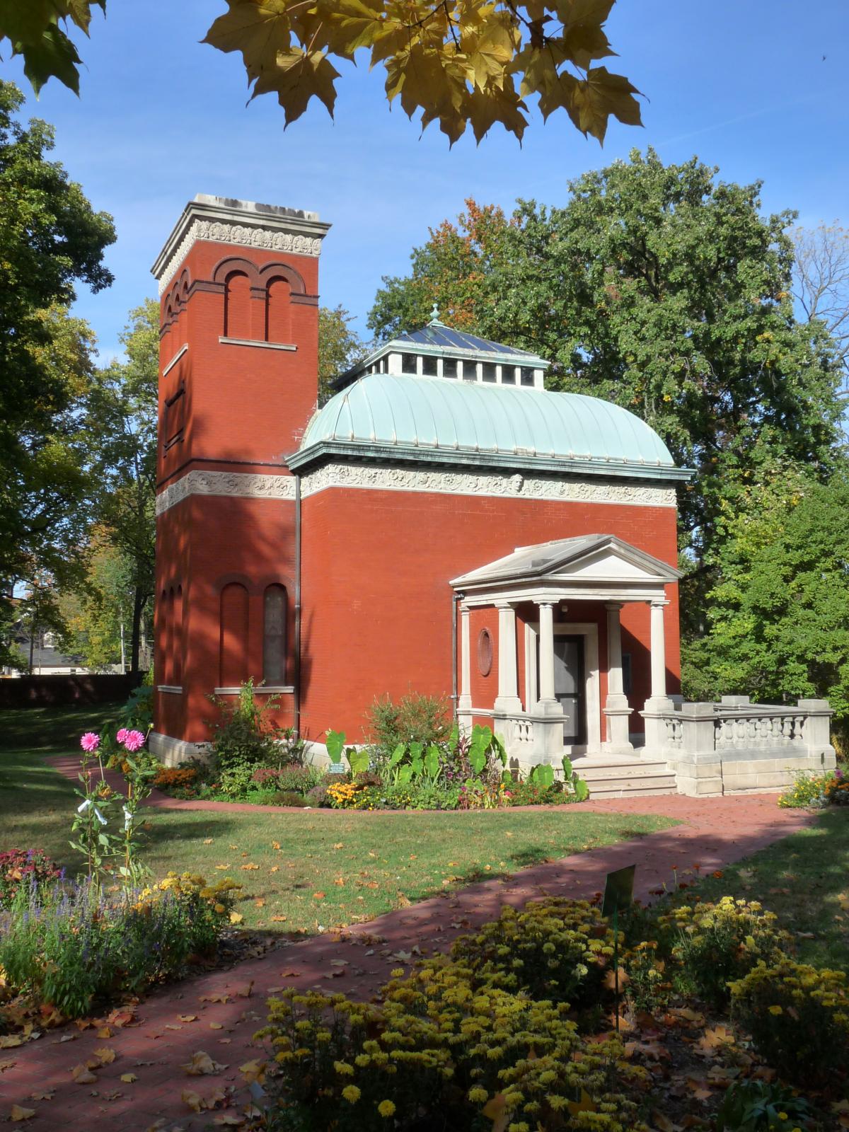 Red brick building with a teal roof and white porch columns, surrounded by green gardens and trees