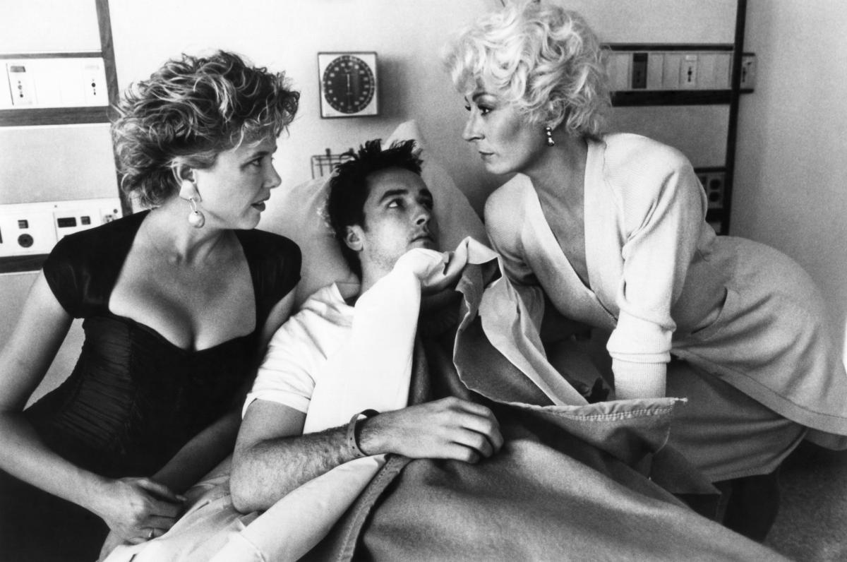 Two women stare each other down while standing over a man in a hospital bed, who looks at the woman on the right with wide eyes