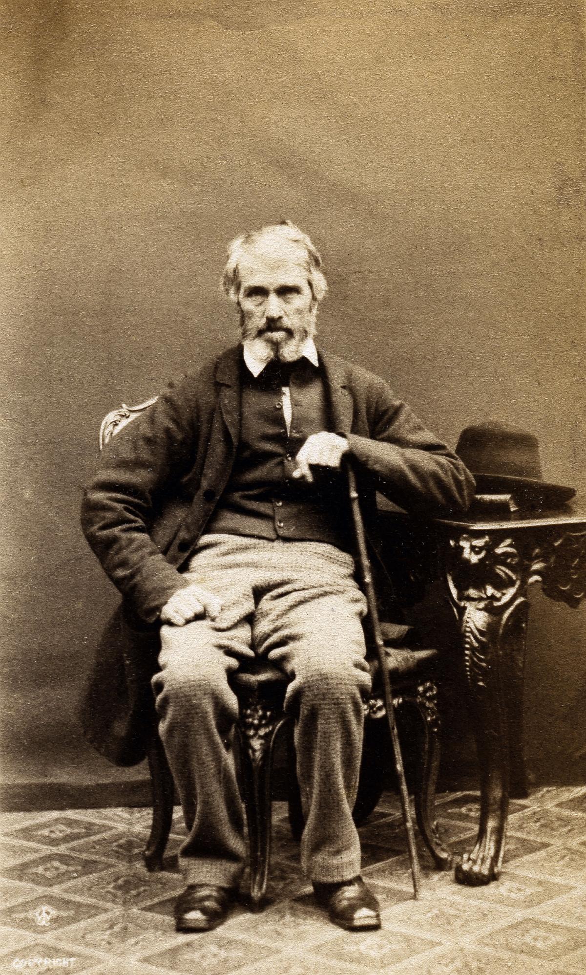 Carlyle sits, resting his left forearm on a walking stick, dressed in grey trousers and a dark coat, his hat sitting on a side table on his left