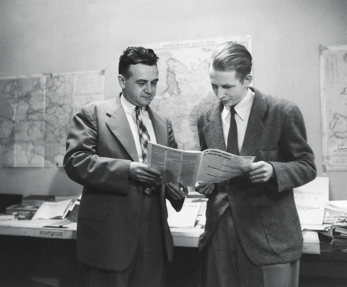 The two men look over a newspaper together, in an office hung with maps