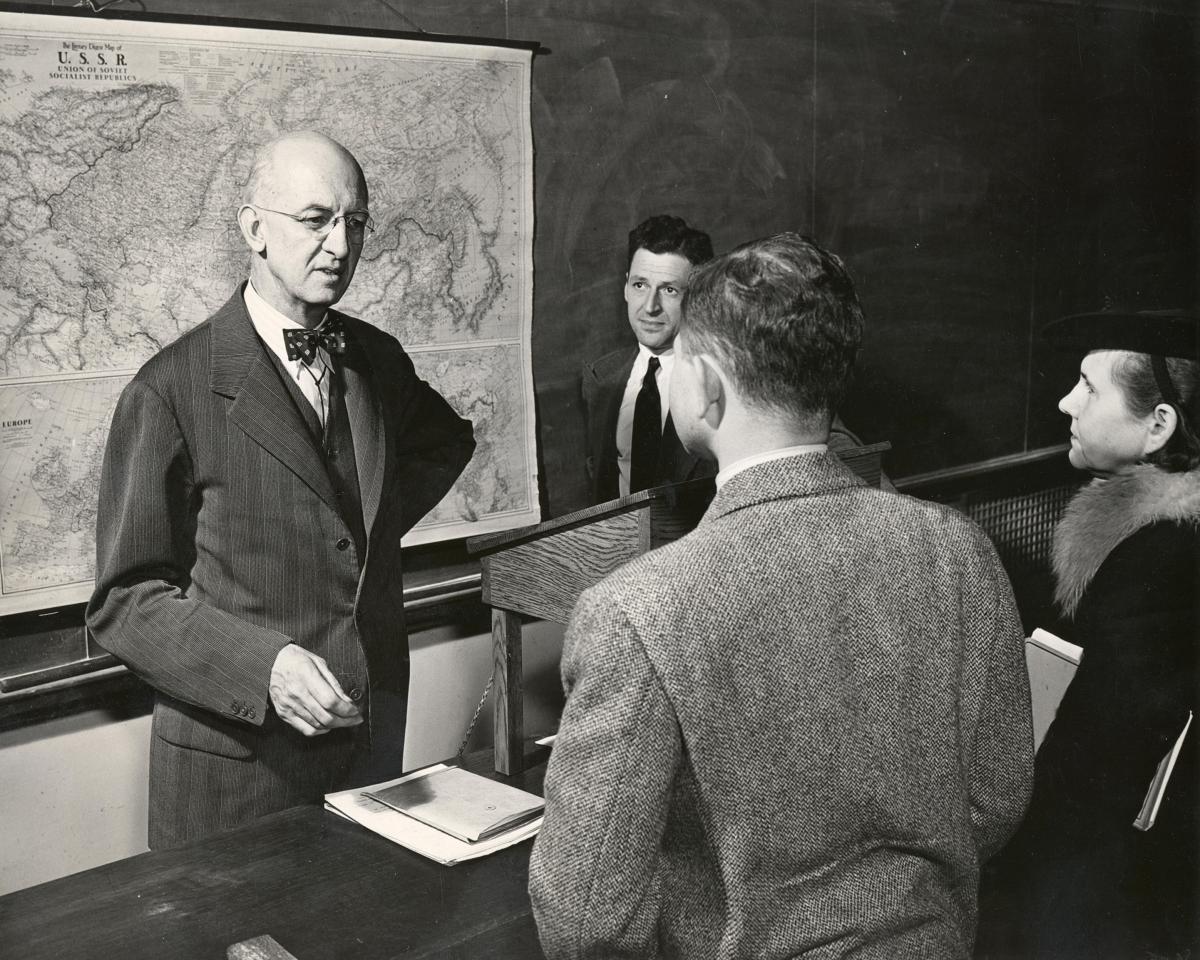 Robinson speaks to two students, his back to a large map and blackboard, while Mosely stands off to his back left