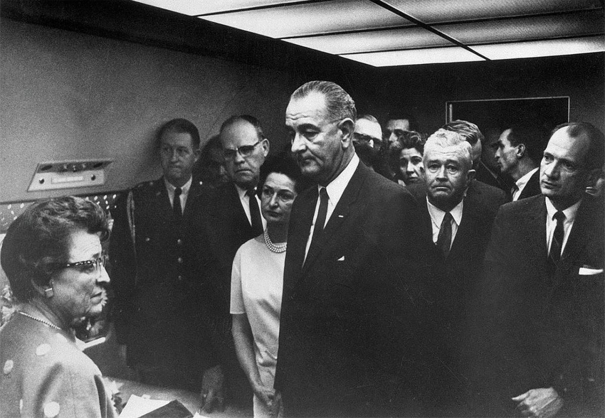 LBJ waits, with a crowd of advisors, including Thornberry, standing behind him