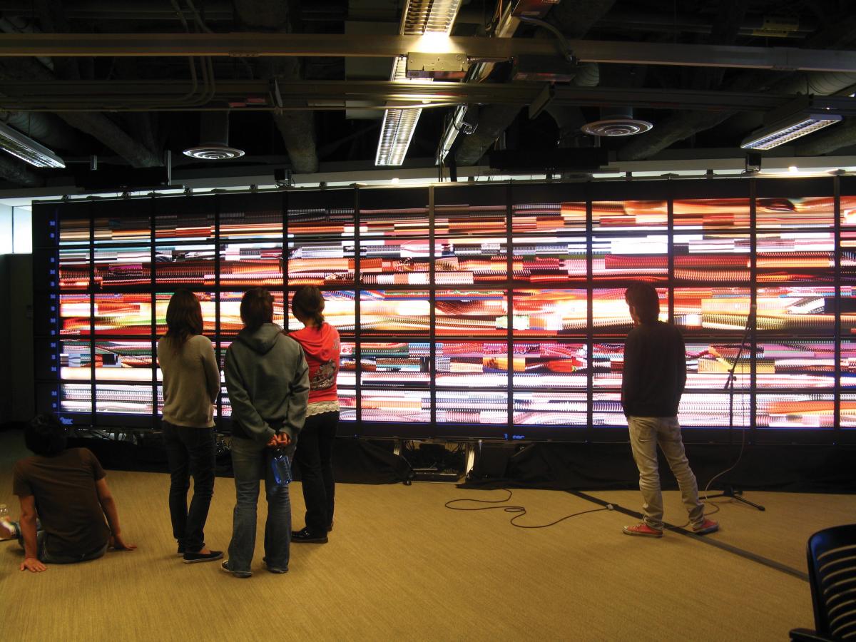 Several people stand in front of numerous small screens set up on a wall