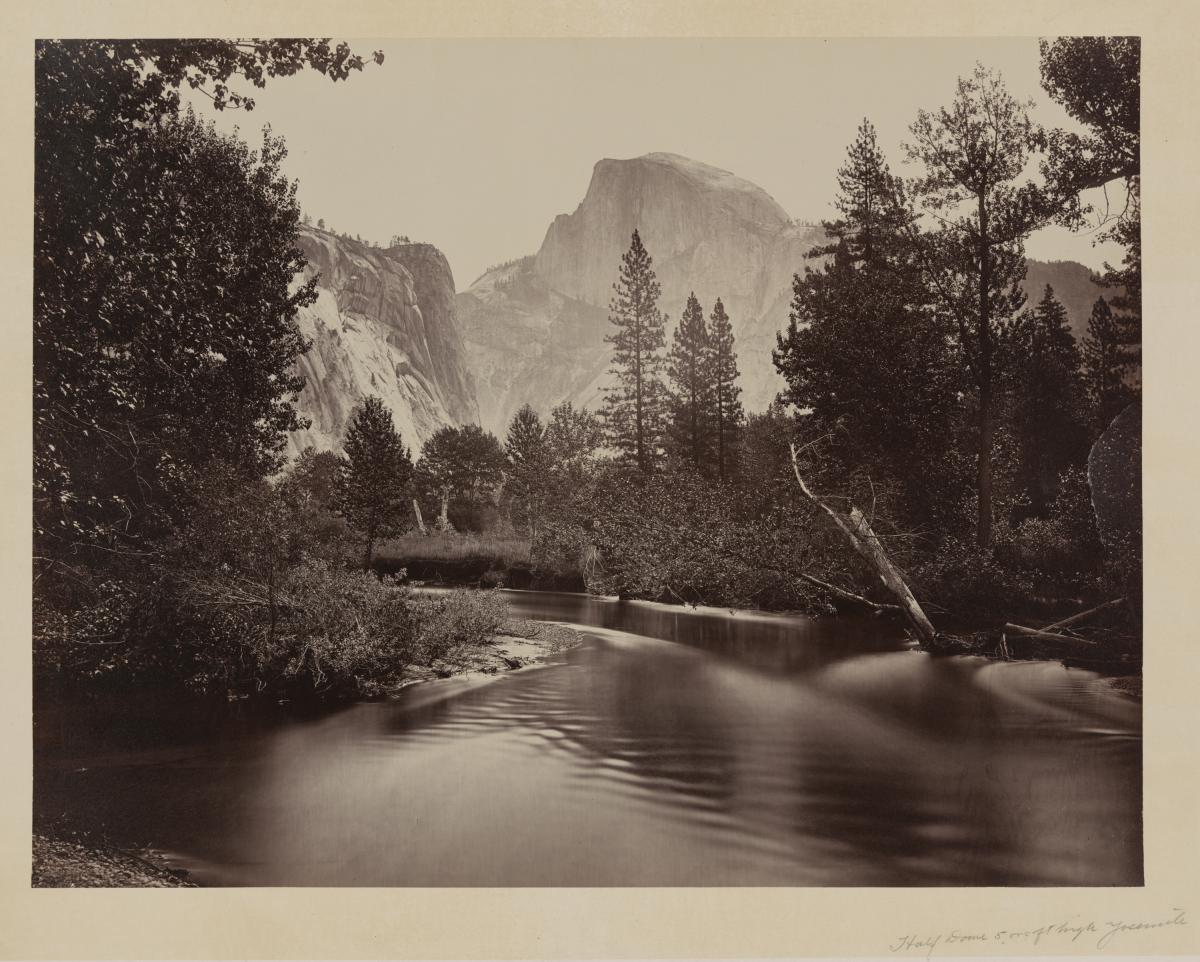 Half Dome, another large rock formation, in the distance, as the backdrop of a calm river and trees