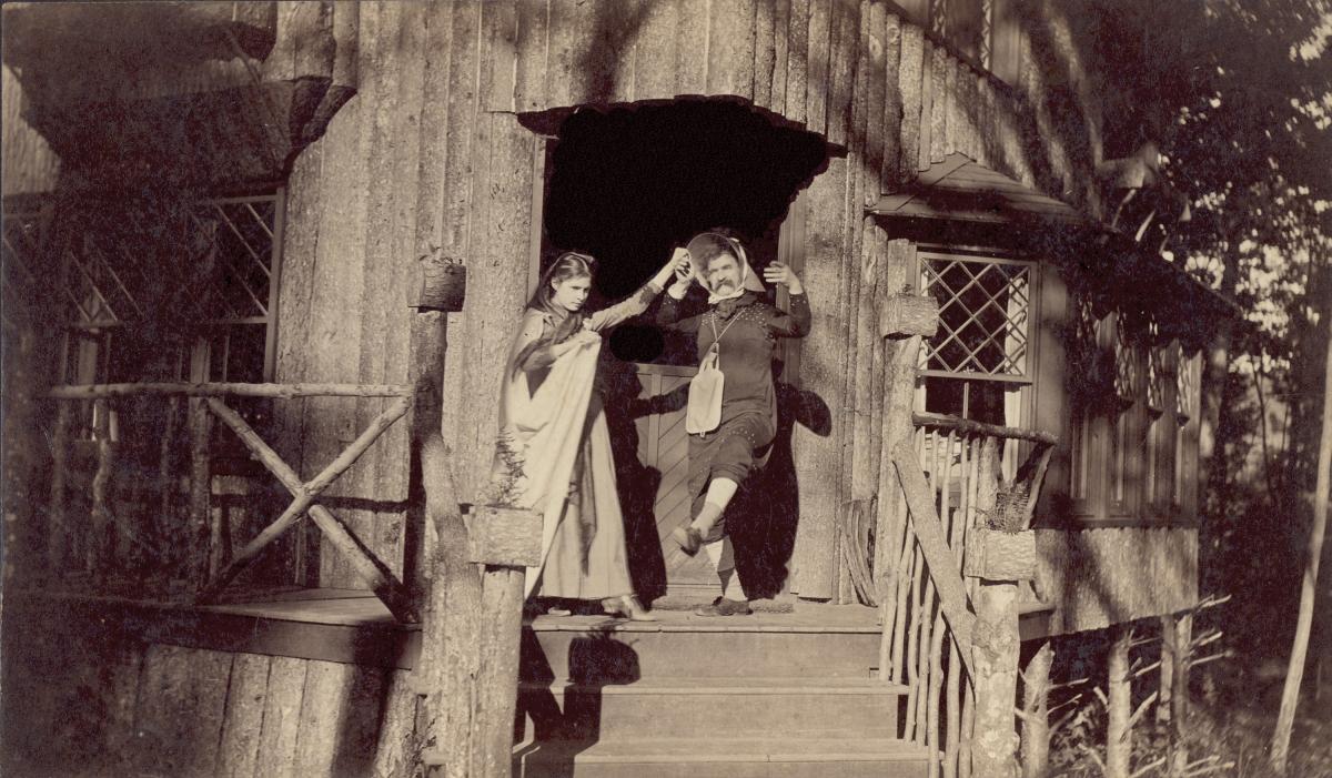 Clemens, with leg outstretched, standing next to Susy on the porch of a wooden house