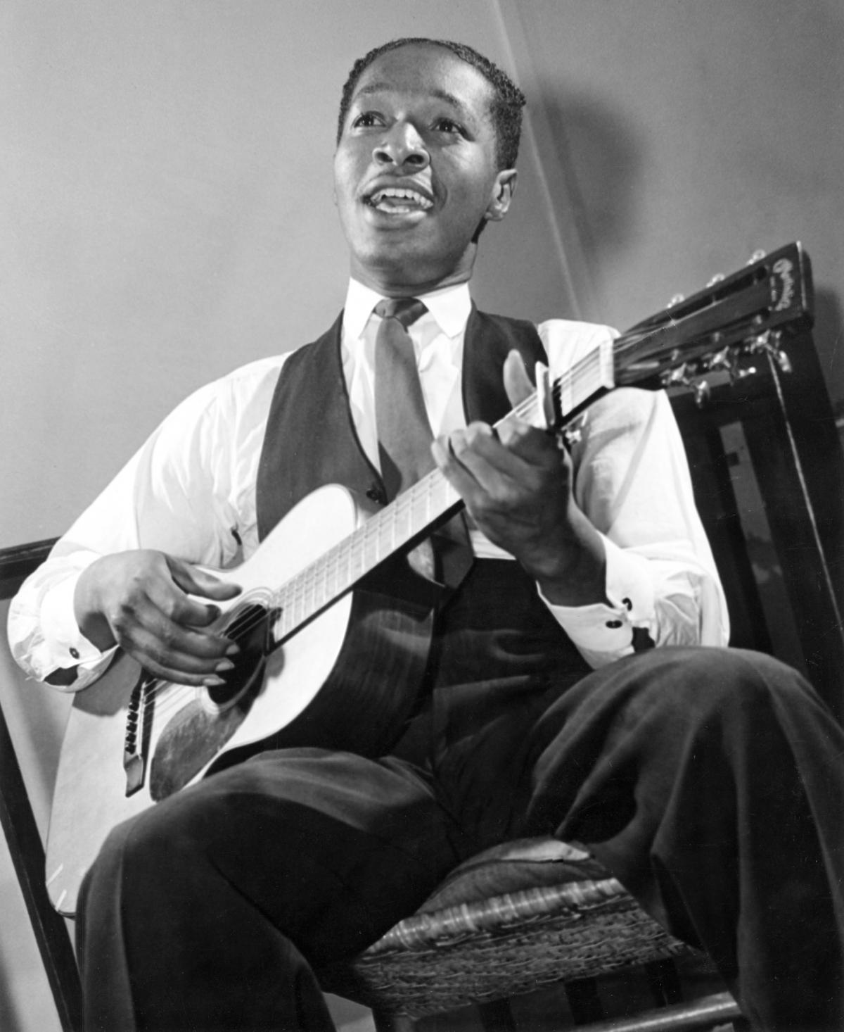 White, wearing a tie and vest, seated, strumming a guitar