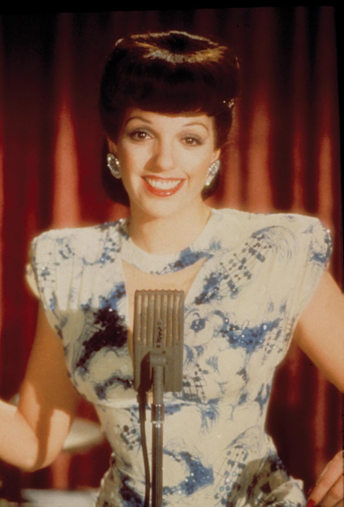 Minnelli with a beehive hairdo, wearing a blue, flowered dress, in front of a microphone, red curtain in the background