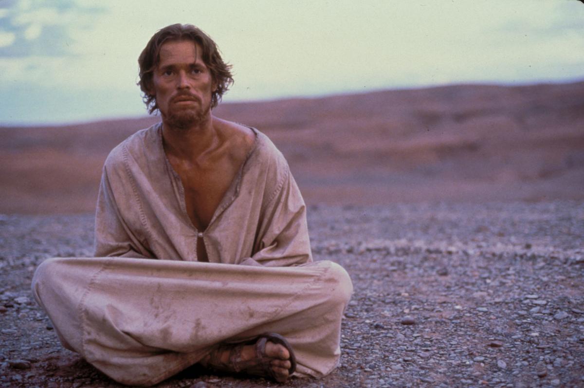 Dafoe, sitting on barren ground, wrapped in dirty rags