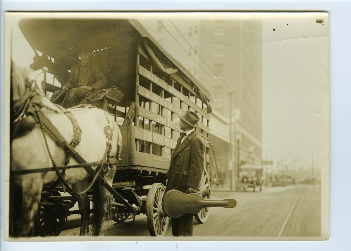 Sandburg, with guitar case in hand, stops to talk to a man driving a horse-drawn wagon, on the street