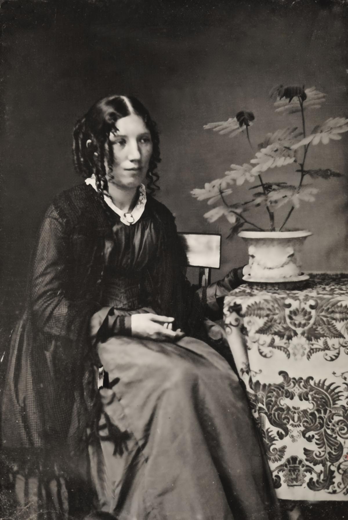 Stowe, with curled hair and wearing a dark shawl and dress, seated next to a table with a flower in a vase