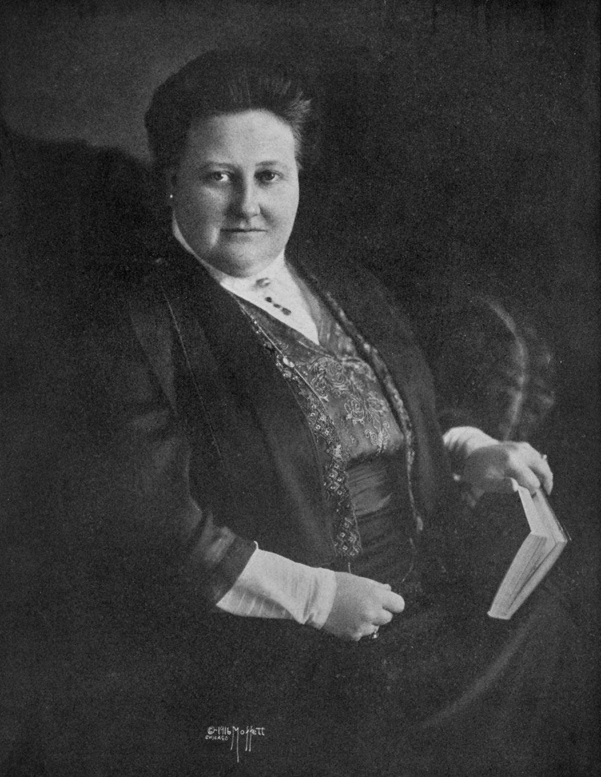 Lowell, wearing a black dress with a high necked white collar, seated