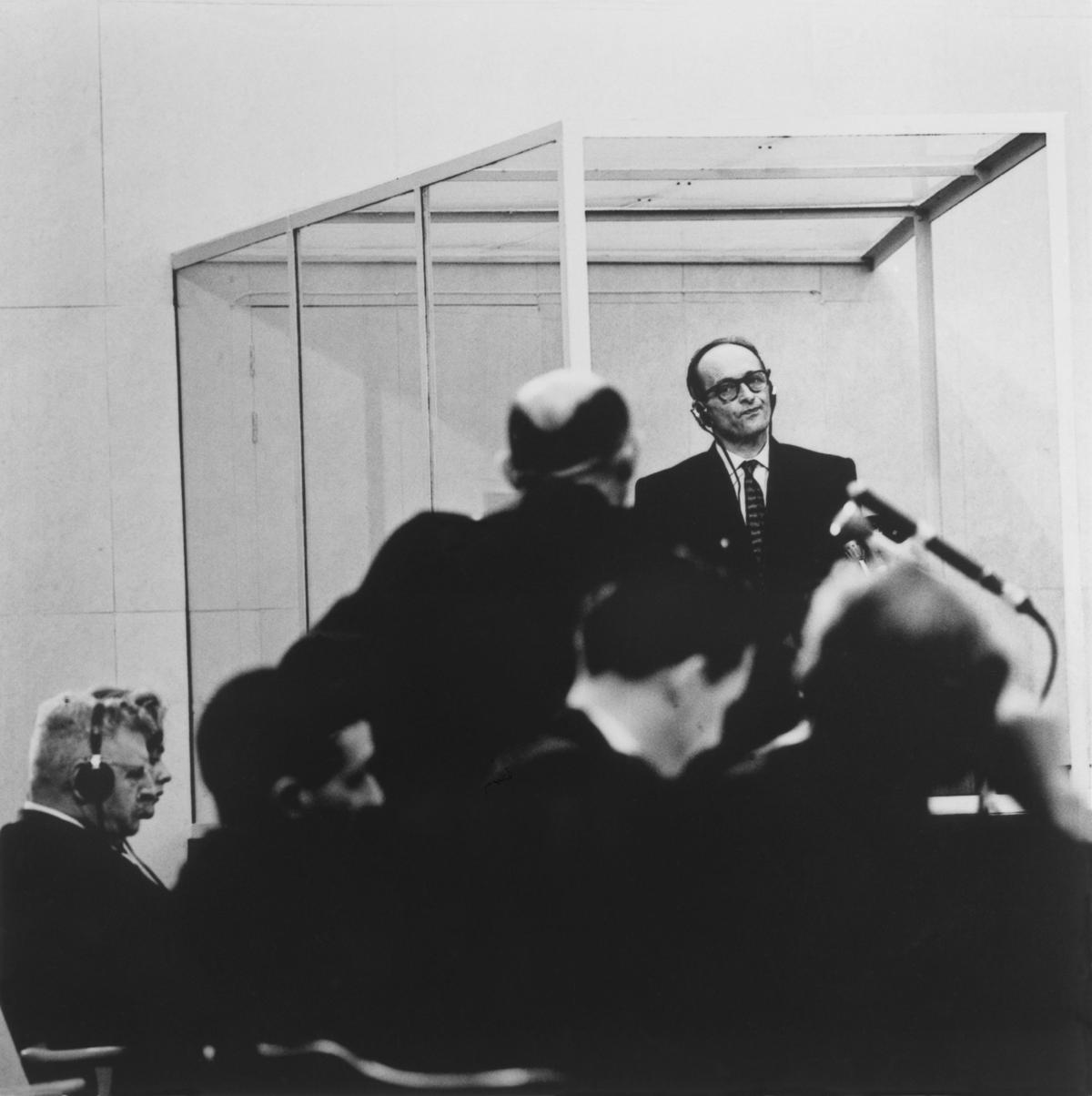Eichmann, wearing glasses, a dark suit, and headphones, purses his lips at a lawyer in the foreground, who is questioning him, hands on his hips