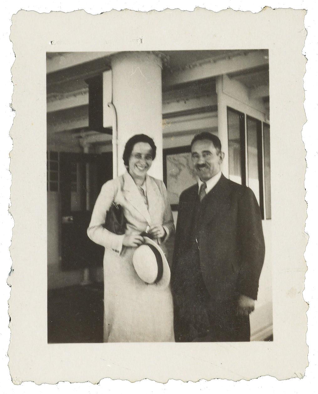 Arendt, smiling and wearing a long white coat, holding a hat, stands next to a moustached man in a dark suit