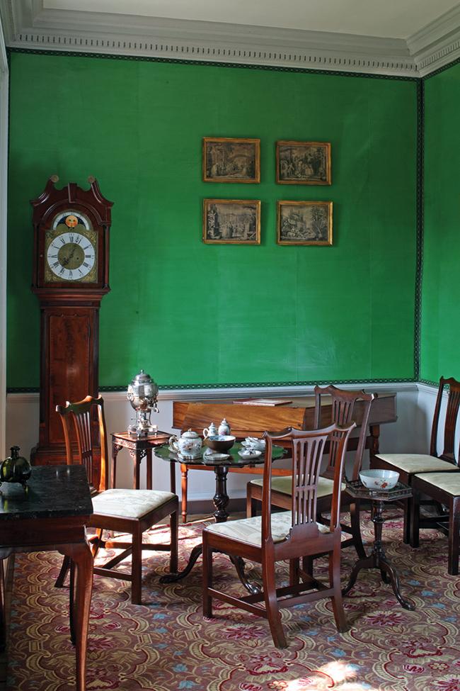 Green walls with crown molding, dark wood grandfather clock in the corner, and several wooden chairs, a coffee table and a desk