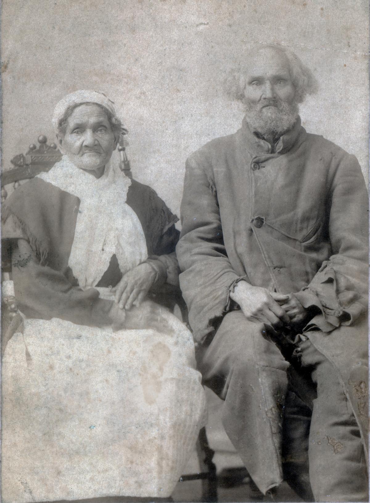 Elderly Hannah and Aaron sit side by side. Hannah in a white lace dress and cap and dark cape, Aaron wearing a gray coat and trousers