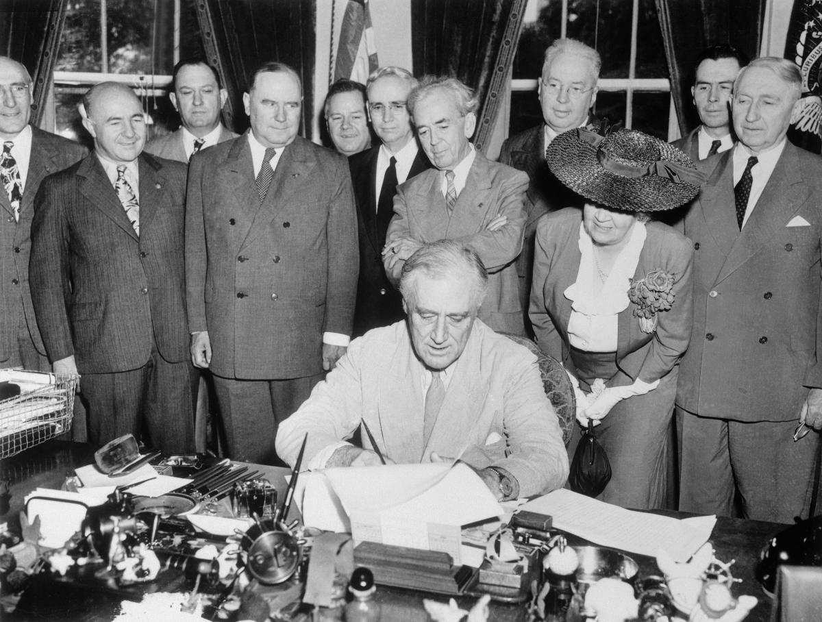 FDR, with his advisors standing behind him, signs the G.I Bill at his desk in the oval office