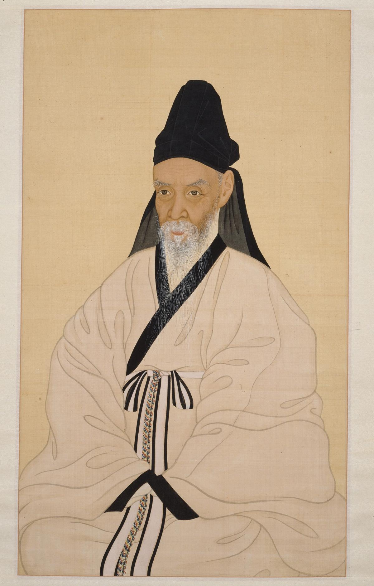 Yi Jae in a white robe, seated with hands folded in his lap, wearing a black cap