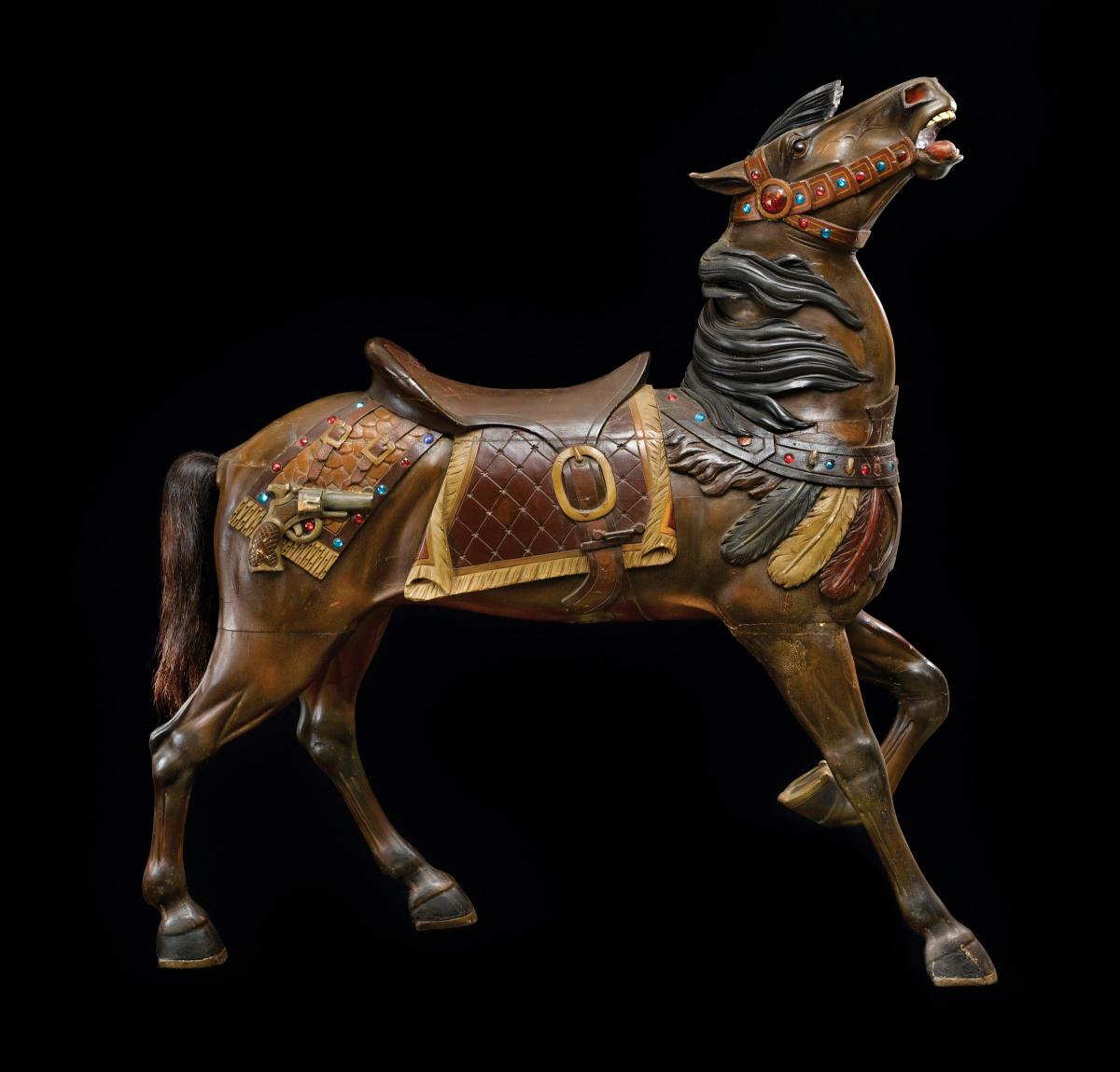 A wooden carousel horse with a raised head, made by Charles Carmel. Done in brown wood, wearing an elaborate saddle and halter