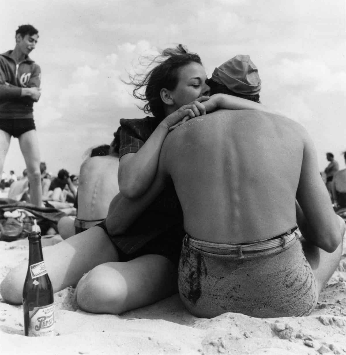 Black and white photo of a man and woman embracing on the beach at Coney Island, with the man's back to the viewer and the woman whispering in his ear